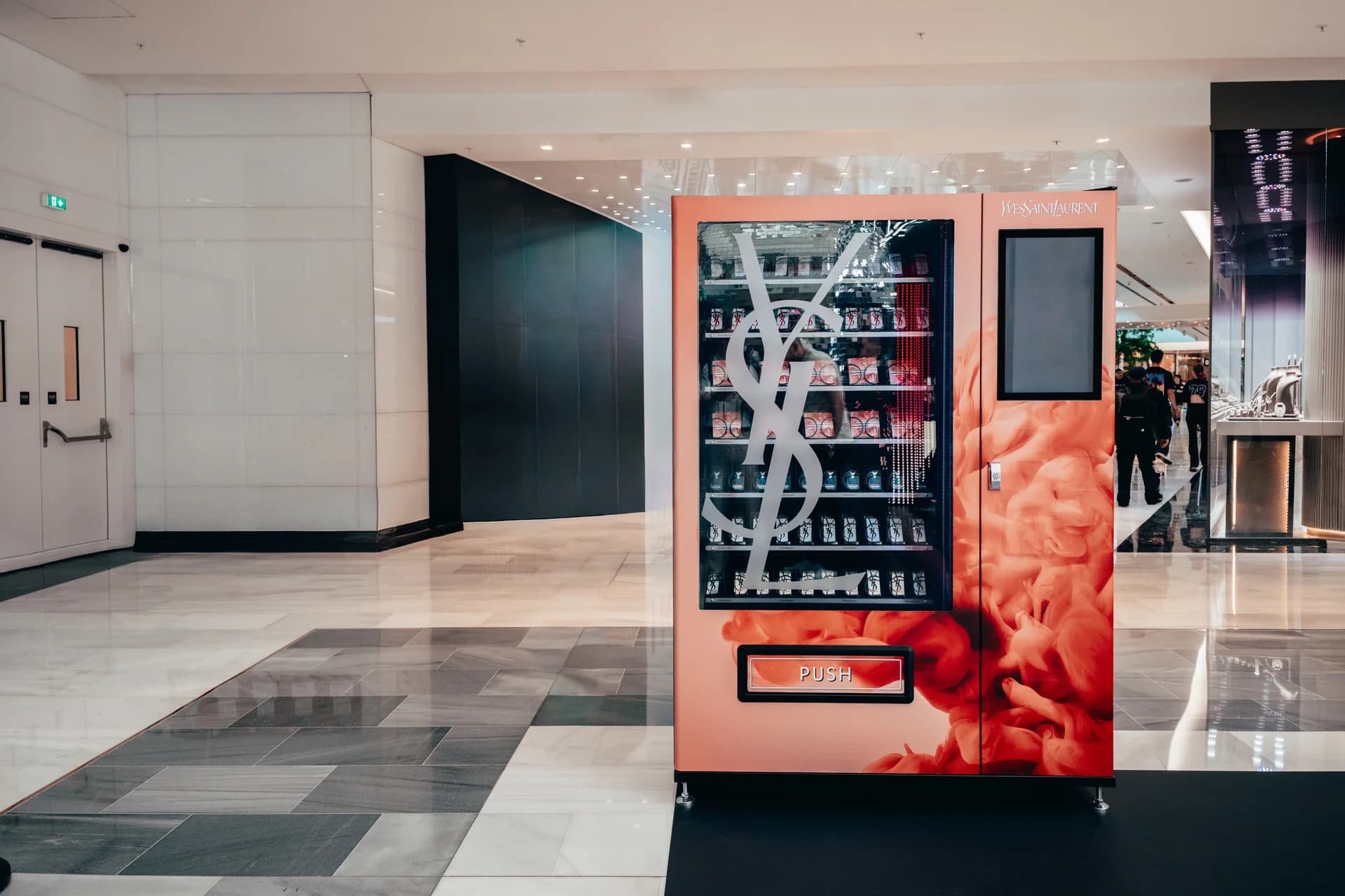 Korálový automat Yves Saint Laurent s velkým monogramem na skle v interiéru obchodního centra.